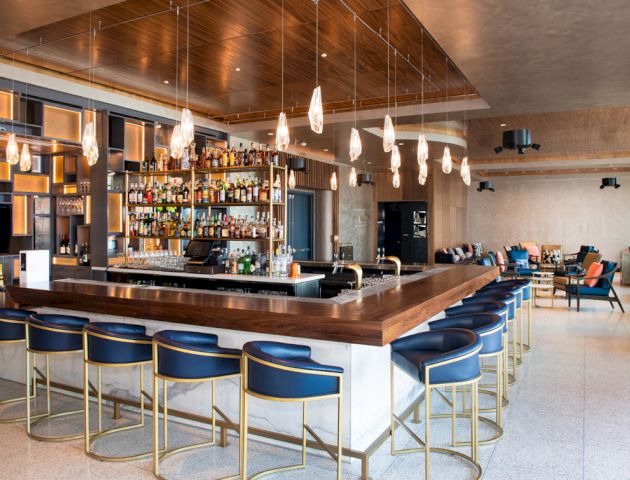 A modern bar with a long counter topped with barstools, pendant lights above, a well-stocked liquor shelf, and a cozy seating area in the background.