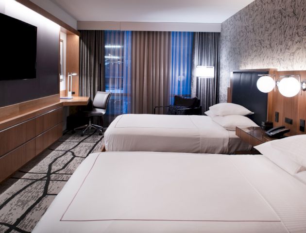 The image shows a modern hotel room with two beds, a flat-screen TV, a desk, a chair, and ambient lighting, designed with contemporary decor.