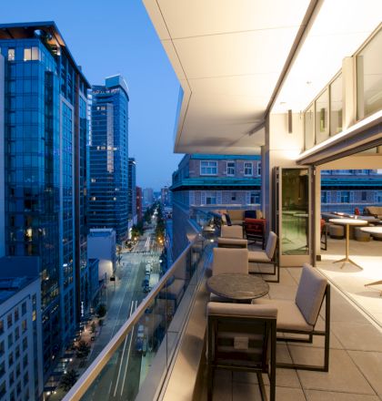 A modern balcony with chairs and tables overlooks a cityscape of tall buildings during twilight, showcasing a mix of glass and concrete structures.