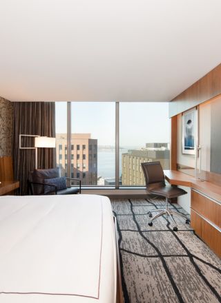 A modern hotel room with a large bed, a wall-mounted TV, a desk with a chair, and a window with a city view and water in the background.