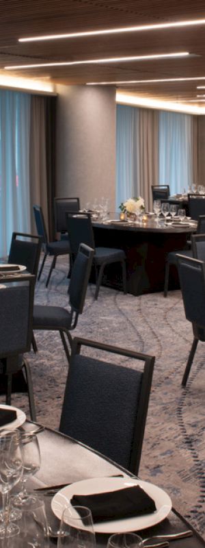 A banquet hall with round tables set for a formal event, featuring dark chairs and tablecloths, under a modern ceiling with recessed lighting.