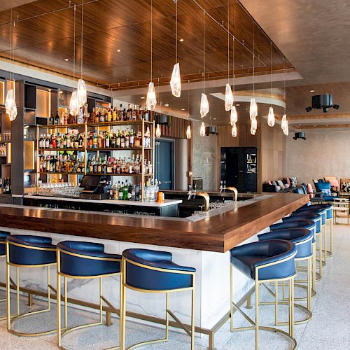 This image shows a modern bar with a wooden countertop, numerous bottles on shelves, hanging lights, and blue bar stools arranged around it.