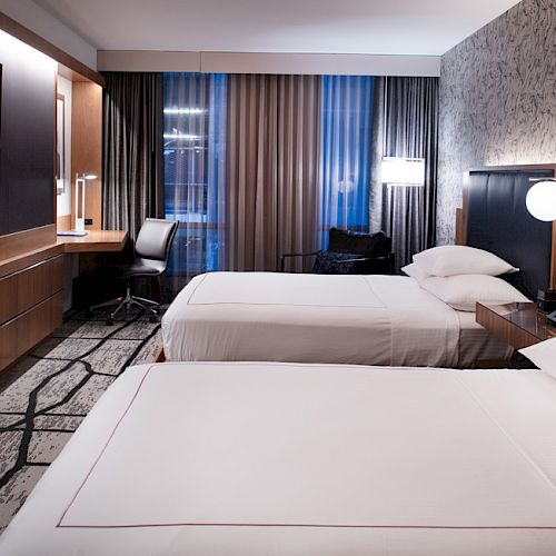 A modern hotel room with two beds, a large TV, a workdesk, lamps, and decorative wallpaper. The room is well-lit with contemporary design.