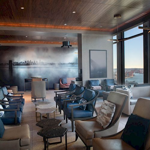 A modern lounge with multiple chairs, tables, large windows, and a mural depicting a city skyline, featuring a view of a ferris wheel and waterfront.