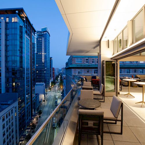 The image shows a modern cityscape from a building's balcony at dusk, with outdoor seating and tables on the balcony, and tall buildings in the background.
