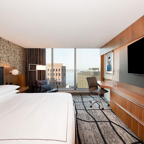 The image shows a modern hotel room with a large bed, a desk, a chair, a flat-screen TV, and a window with a city view.