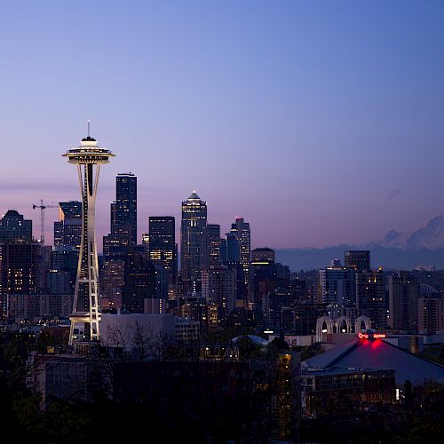 The image showcases the Seattle skyline at dusk, featuring the Space Needle prominently, with Mount Rainier visible in the background.