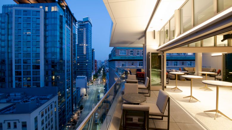 This image shows a modern cityscape at dusk with tall buildings, and a well-lit rooftop terrace featuring tables, chairs, and large glass windows.