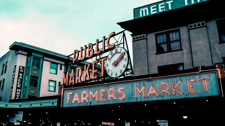 The image shows a public market with a prominent "Farmers Market" neon sign and clock. There are buildings in the background, one with a sign reading "MEET THE...".