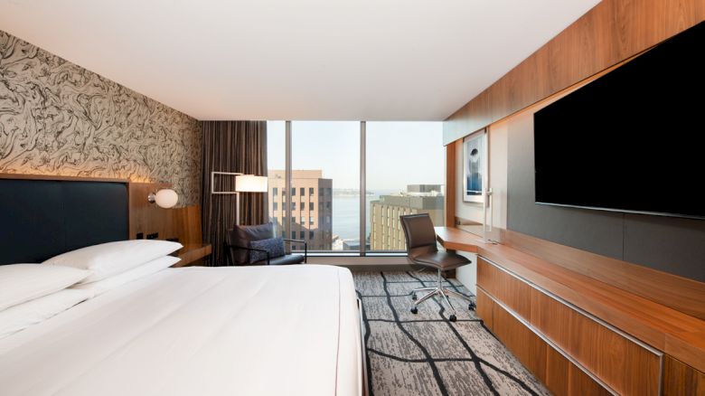 A modern hotel room with a large bed, wall-mounted TV, desk, and chair, offering a view of buildings and the water through large windows.