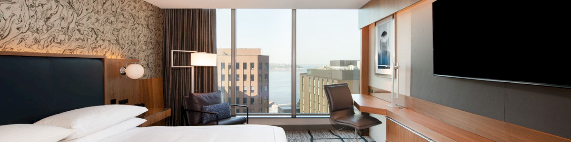 Modern hotel room with a large bed, desk, chair, and TV. Features floor-to-ceiling windows with a city and water view.