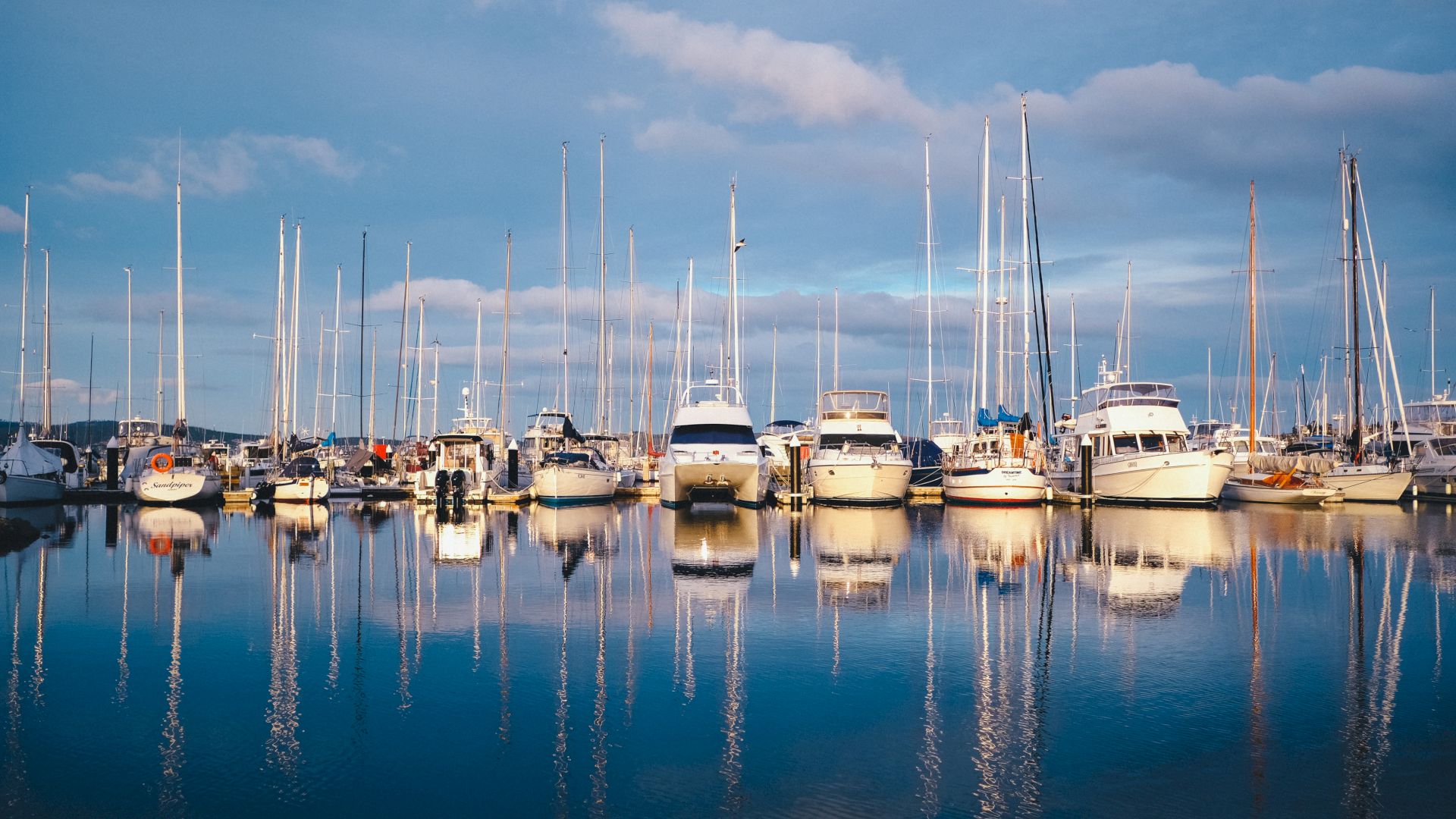 A serene marina with several boats and yachts docked, their masts reflecting on the calm water under a partly cloudy sky, creating a picturesque scene.