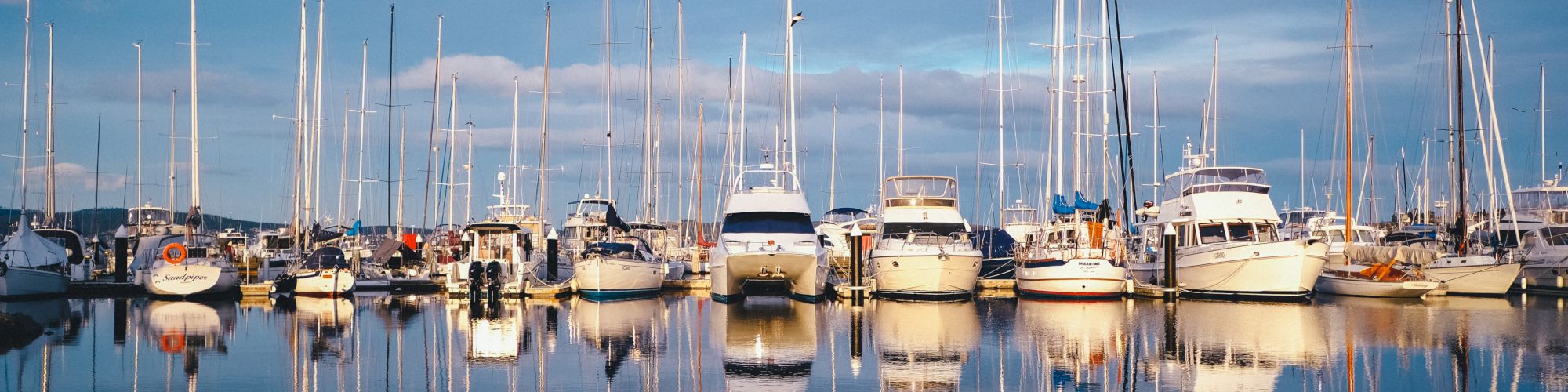 A serene marina with several boats and yachts docked, their masts reflecting on the calm water under a partly cloudy sky, creating a picturesque scene.