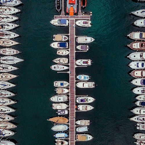 An aerial view of numerous boats docked along a narrow pier extending into the water. The water surrounding the boats is a deep blue color.